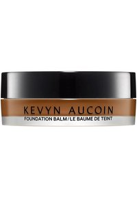 Foundation balm in a round jar, featuring a black lid and a beige-brown body with clear base. Text includes "Kevyn Aucoin" and product name.