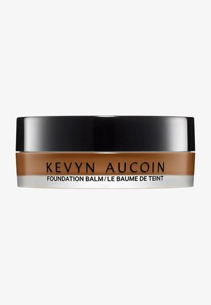 Balsamo fondotinta in un barattolo rotondo, con un coperchio nero e un corpo beige-marrone con base trasparente. Il testo include "Kevyn Aucoin" e il nome del prodotto.