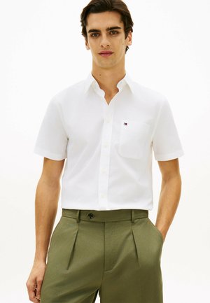 Ung mand iført en kortærmet hvid skjorte med knapper og brystlomme samt olivengrønne plisserede bukser, stående foran en ensfarvet baggrund.