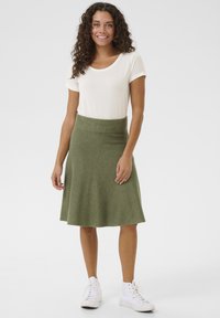 Jupe verte, longueur genou, avec une taille ajustée et un design évasé, associée à un haut blanc à manches courtes et des baskets blanches.