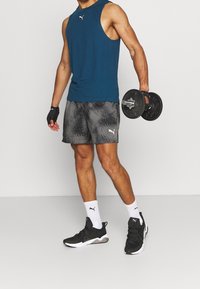 Teal Puma tanktop, grijze gestippelde shorts, zwarte sportschoenen. Houdt een dumbbell vast, draagt polssteunen en witte sokken.