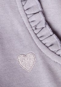 Jasnofioletowa dzianina z falbanką na brzegu oraz małe haftowane serce w kontrastującym kolorze nici w pobliżu dekoltu.