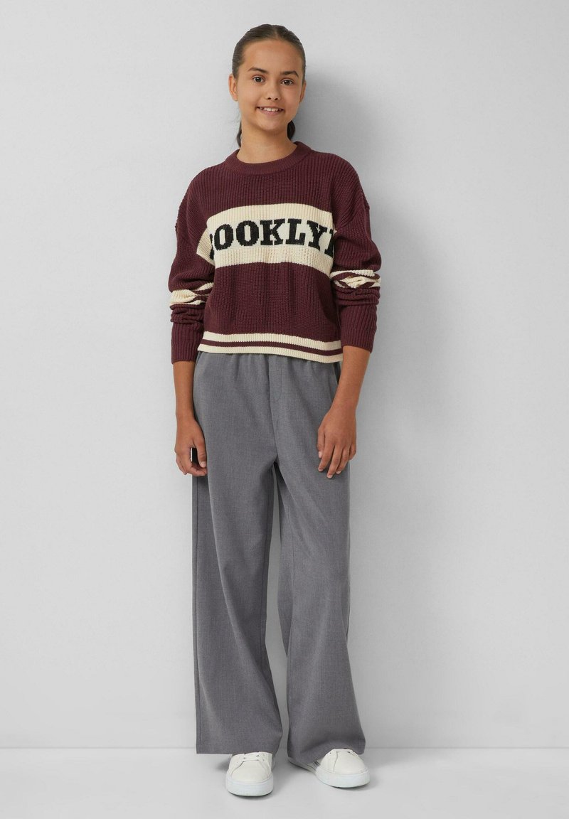 Bordeaux gebreide trui met "BROOKLYN" in het zwart, gecombineerd met lichtgrijze wijde broek en witte sneakers.