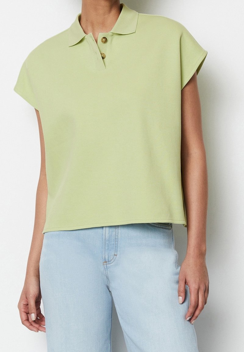 Haut polo vert clair avec un col, des manches courtes en cape, une patte de boutonnage à trois boutons et une coupe décontractée. Assorti avec un jean bleu clair.