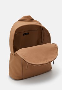 Tan ryggsäck i texturerat material, med ett generöst utrymme, en framficka och ett zip-eller fack inuti. Försedd med en guldfärgad dragkedja.