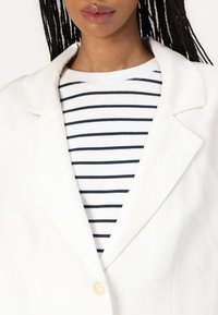 Hvit skreddersydd blazer med spisse revers, enkel knappelukking, lagd over en marineblå og hvit stripete skjorte. Glatt tekstur.