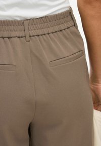 Hnědé, strukturované látkové kalhoty s elastickým pasem a dvěma zadními kapsami. Design se vyznačuje jemným prošíváním a volným střihem.