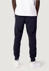 Persona che indossa pantaloni jogger neri, sneakers bianche e una maglietta bianca a maniche corte, in piedi di spalle contro uno sfondo semplice.