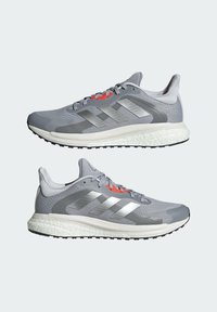 Paire de chaussures de course grises et argentées avec des accents orange sur les lacets et des semelles blanches rembourrées, vues de côté sur un fond clair.