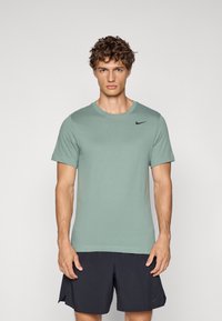Camiseta de manga corta verde claro hecha de tela suave, con un logotipo negro de Nike en la parte superior izquierda del pecho. Combinada con pantalones cortos oscuros.