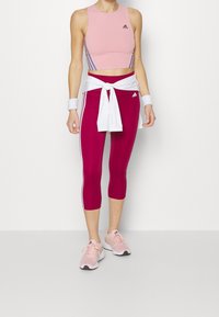 Lyserødt træningstop med blå striber, parret med maroon capribukser med hvide sidestumper. Hvid skjorte knyttet foran og lyserøde sneakers.