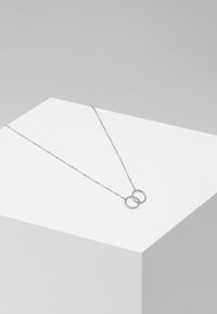 Collana in metallo argentato con due pendenti circolari intrecciati, appesa a una delicata catena, esposta su una superficie bianca e angolare.