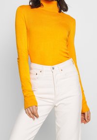 Jasnopomarańczowy sweter typu golf wyk wykonany z miękkiego materiału, połączony z białymi dżinsami o wysokim stanie. Rękawy mają ściągacze z prążkowanego materiału.