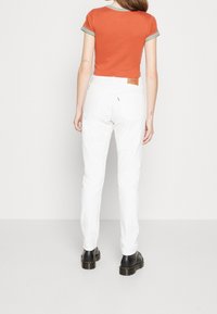 Pantalones de mezclilla blancos con corte recto, que presentan un parche de cuero en la cinturilla trasera; combinados con una blusa naranja de manga corta con ribete a rayas.