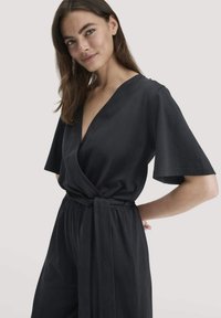 Frau mit langen braunen Haaren, die ein schwarzes Wickelkleid mit kurzen Ärmeln und einem gebundenen Taillengürtel trägt, lächelt sanft.