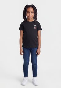 Κορίτσι με κοτσίδες που φοράει μαύρο T-shirt με γραφικά, μπλε τζιν και λευκά αθλητικά παπούτσια, στέκεται μπροστά σε απλό φωτεινό φόντο.