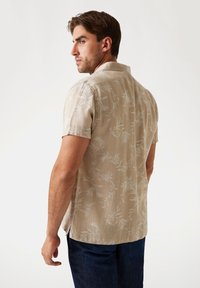 Camisa de manga corta, beige, con un patrón botánico y un cuello muesca. Fabricada en tela ligera, presenta aberturas laterales para mayor comodidad de movimiento.