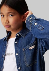 Camicia di jeans blu con bottoni bianchi, una tasca sul petto e etichetta "Calvin Klein". Sotto, una maglietta bianca con dettagli del logo argentati.