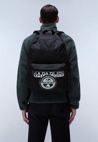 Zaino in tessuto nero con una grande tasca frontale, caratterizzato da un logo circolare e testo. Il design include spallacci regolabili e una texture morbida.