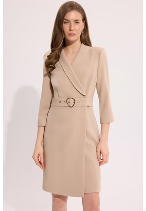 Kobieta z długimi falującymi włosami, ubrana w beżową sukienkę owijaną z paskiem i rękawami do połowy przedramienia, na jednolitym białym tle.