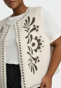 Personne portant un gilet en polaire blanc avec une broderie florale marron foncé, par-dessus une chemise blanche à manches courtes et un pantalon noir, montrée de la poitrine aux hanches.
