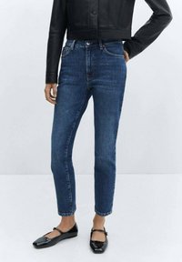 Jean en denim bleu foncé à coupe fuselée, avec une taille mi-haute, cinq poches et une fermeture à bouton. Associé à des ballerines noires.