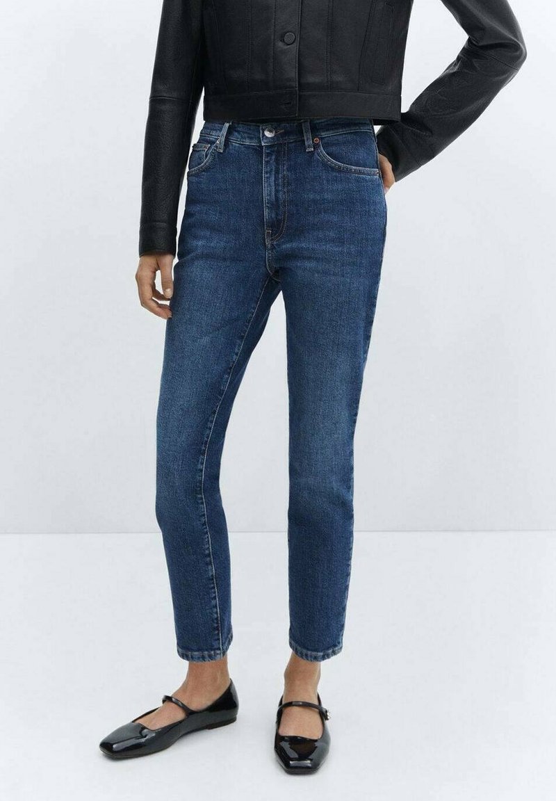 Jean en denim bleu foncé à coupe fuselée, avec une taille mi-haute, cinq poches et une fermeture à bouton. Associé à des ballerines noires.