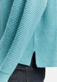 Türkisfarbener, gerippter Pullover mit seitlichem Schlitz, aus strukturiertem Strickstoff, über dunklen Denim-Jeans getragen.