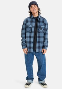 Camisa de franela a cuadros azul y marino con dos bolsillos en el pecho, llevada sobre una camiseta negra, combinada con unos vaqueros de mezclilla azules holgados y zapatillas negras.