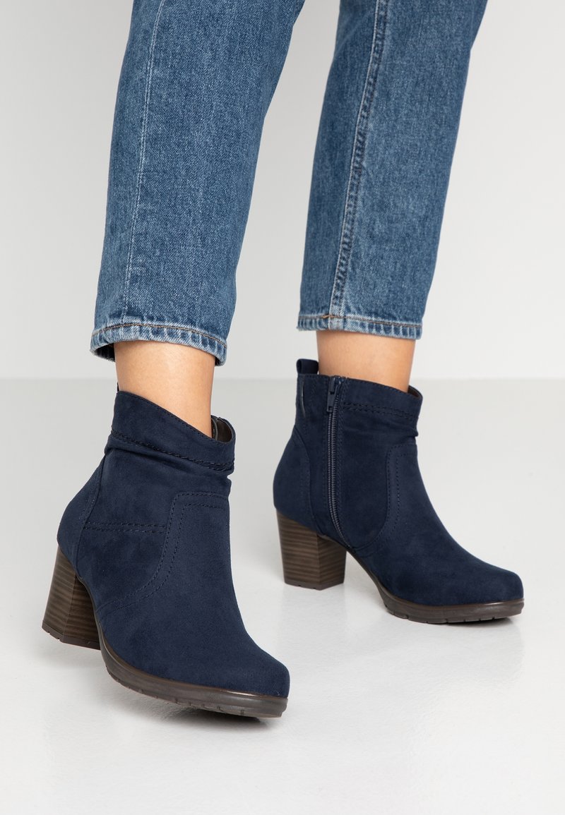 Personne portant des bottines en daim bleu foncé à talons blocs et un jean bleu raccourci, debout sur un sol blanc.