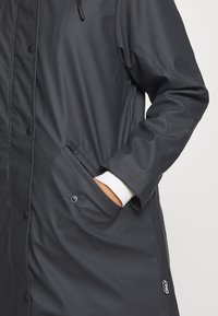 Lang svart vanntett jakke med glidelåsdesign, trykknapper, sidelommer og ribbestrikkede ermer. Glatt, mat overflate med logo.