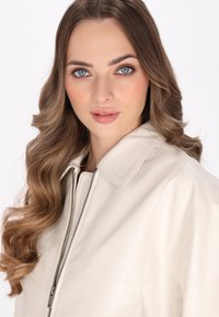 Lichtbeige leren jas met een kraag, met een rits aan de voorkant en een gladde textuur. Haar gestyled in zachte golven, vastgelegd in een close-up opname.