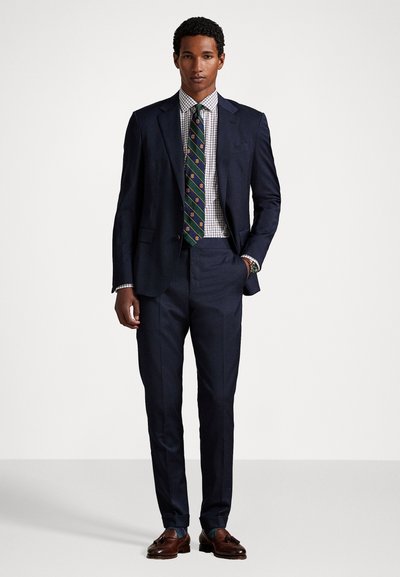 Traje azul marino con una americana ajustada y pantalones a medida, combinado con una camisa de cuadros y una corbata de estampado. Zapatos de cuero marrón.