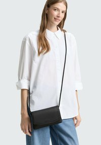 Borsa a tracolla in pelle nera con un design semplice, forma rettangolare e texture liscia, indossata sopra una camicia bianca a bottoni e pantaloni in denim.