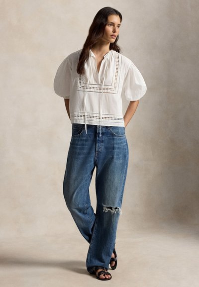 Γυναίκα που φοράει λευκό μπλουζάκι με φουσκωτά μανίκια και δαντελένιες λεπτομέρειες, καθώς και άνετα μπλε τζιν με σκίσιμο στο γόνατο, στέκεται με τα χέρια πίσω από την πλάτη και κοιτάζει πλάγια.