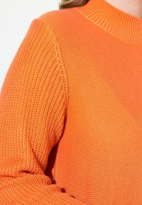 Oranssi neulepusero, jossa on vaihtelevaa tekstuuria; ribattu kaula-aukko ja hihnat yhdistyvät sileään kroppaan. Pehmeä kangas ja lämmin sävy.