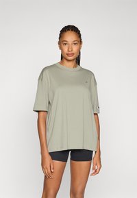 Oliven grønn bomulls T-skjorte med korte ermer, løs passform, rund hals og liten logo-detall på brystet. Brukt over svarte shorts.