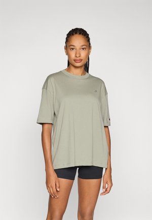 Olive green cotton T-shirt with short sleeves, loose fit, crew neckline, and small logo detail on the chest. Worn over black shorts.