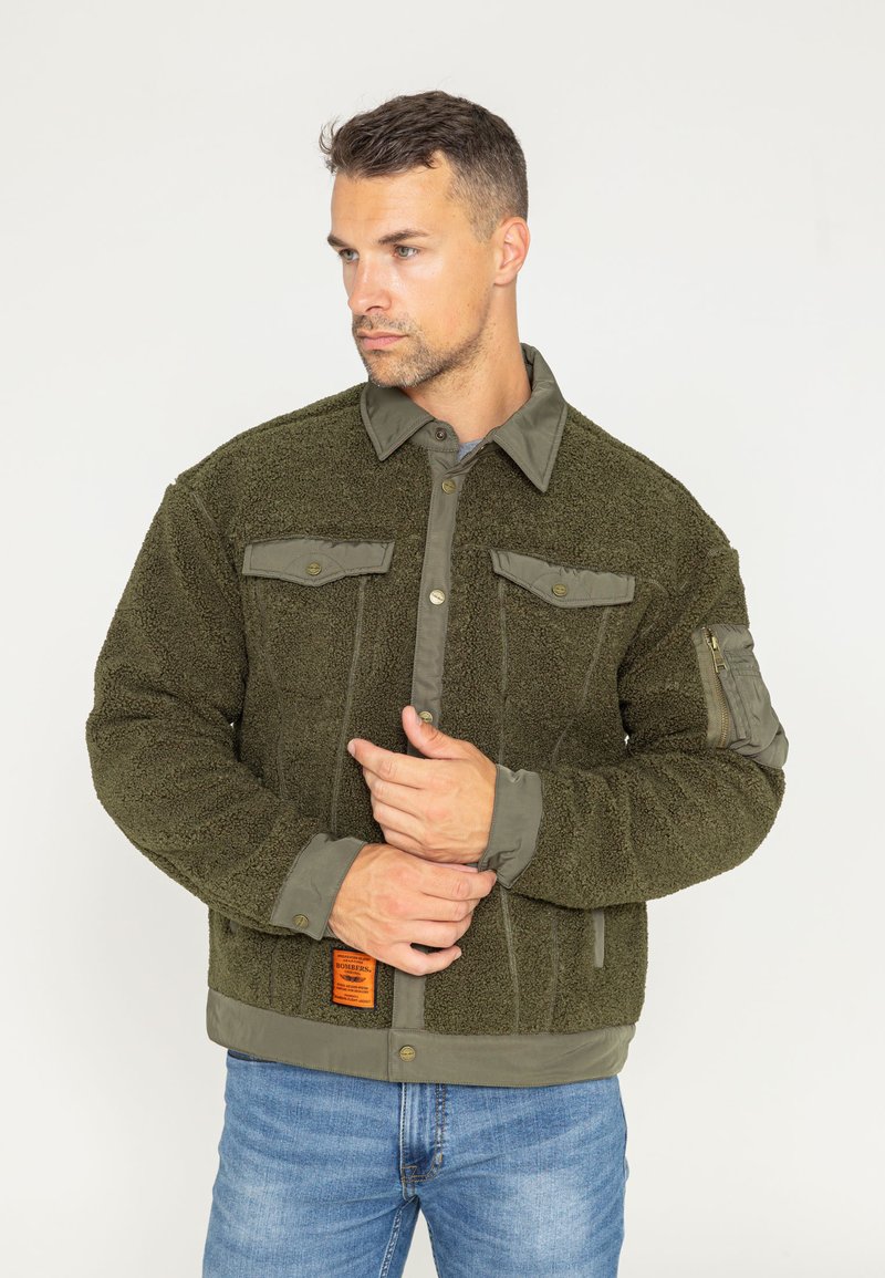 Oliwkowa bluza polarowa o miękkiej strukturze, z kontrastowymi detalami na ramionach i kołnierzu, dwoma kieszeniami na piersi oraz kieszenią na rękawie z zamkiem błyskawicznym.