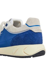 Modré tenisky s textúrovaným semišovým opätkom a hladkým syntetickým zvrškom. Majú krémovú podrážku s čiernymi gumovými akcentmi a jemne polstrovaný jazyk.