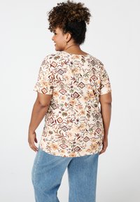 Een bloemenpatroon, korte mouwen shirt in beige met oranje, groen en kastanjebruine accenten, gecombineerd met blauwe jeans. Soepele katoen textuur.