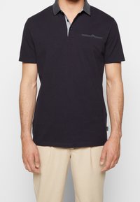 Námořnická modrá polo košile s vzorovaným límcem, knoflíkovou légou a malou kapsičkou na prsou. Vyrobena z měkké tkaniny s krátkými rukávy.