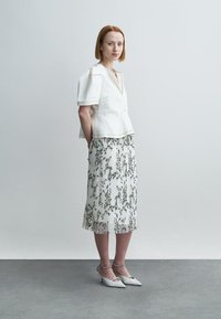 Λευκό τοπ με δομημένη υφή και κοντά μανίκια, σε συνδυασμό με πλισέ φλοράλ φούστα σε λευκό και πράσινο, συμπληρωμένο με λευκά παπούτσια με τακούνι.