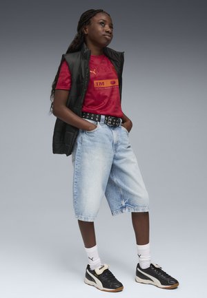 T-shirt rossa Puma con logo, gilet senza maniche nero, pantaloni corti in denim larghi di colore blu chiaro, scarpe da ginnastica bianche e nere, e calzini bianchi con logo.