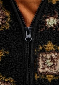 Crna polar jakna s patentnim zatvaračem i teksturiranim uzorkom u smešnoj, žutoj i zelenoj boji. Unutrašnjost je narandžasta. Patentni zatvarač je crn.