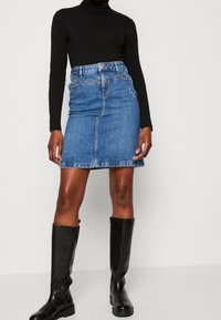 Kvinna i blå denimkjol, svart långärmad polotröja och svarta knähöga stövlar står mot en enfärgad bakgrund.