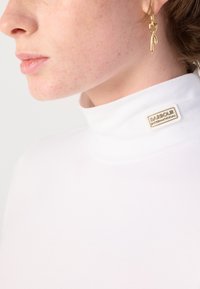 White turtleneck shirt with a ribbed texture and a small cream label reading "Barbour International." Gold hoop earring with a twisted design.