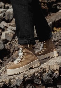 Stivali in suede marrone con un colletto in finta pelliccia bianca texturizzata, dettagli in oro e una sola spessa. Indossati con jeans neri risvoltati su un terreno roccioso.