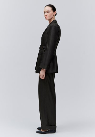 Femme debout de profil portant un blazer noir taillé sur mesure et un pantalon large avec des chaussures plates noires sur un fond clair uni.