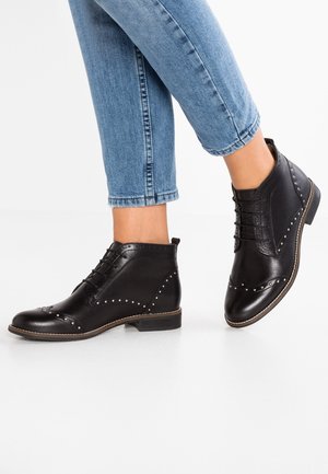 Zwarte leren enkellaarsjes met studs en veters, gecombineerd met cropped lichtblauwe spijkerbroeken tegen een witte achtergrond.